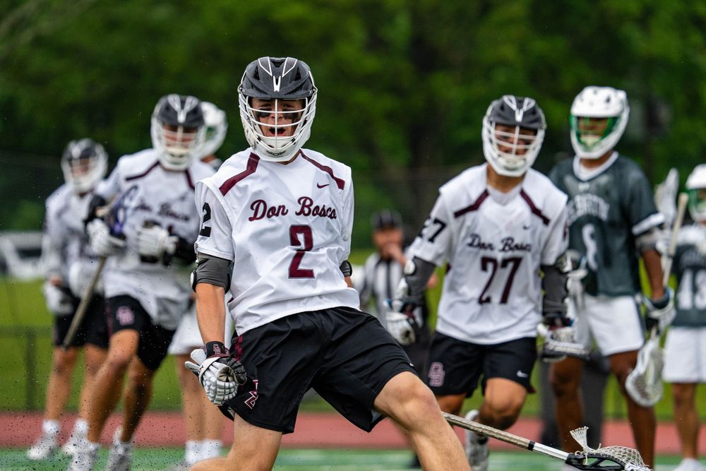 Don Bosco lacrosse players in white uniforms compete on a green field during a youth lacrosse game.