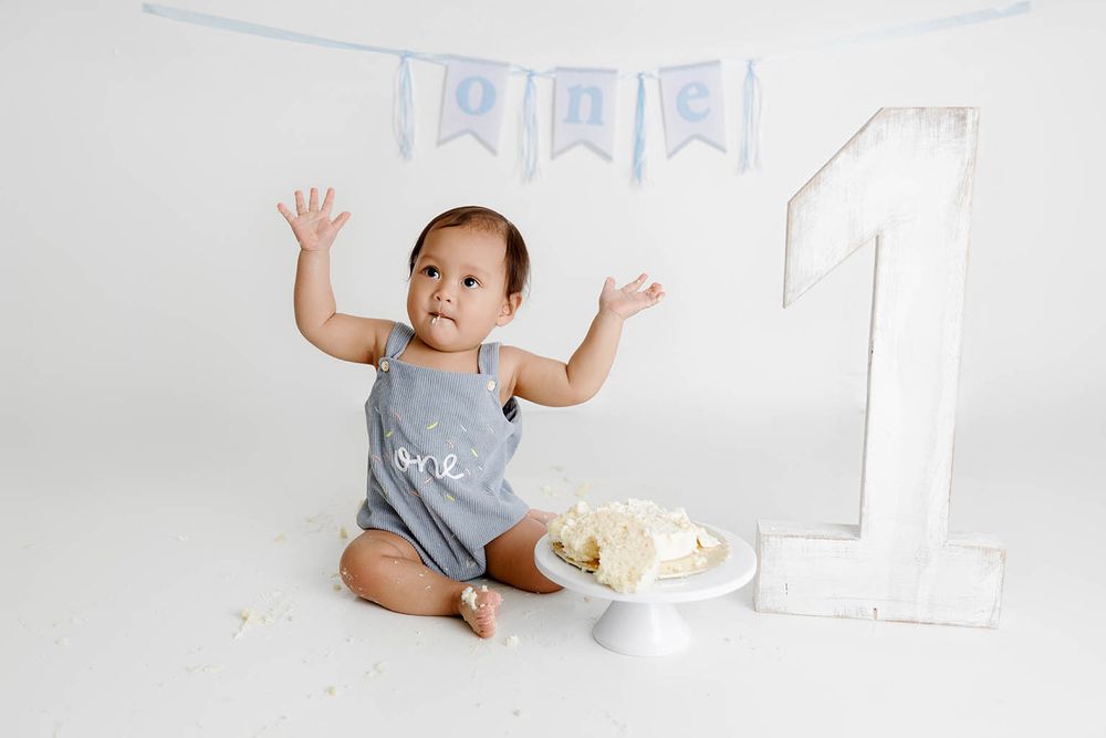 Baby boy in gray romper celebrates first birthday with smash cake, blue ONE banner, and large number 1 decoration.