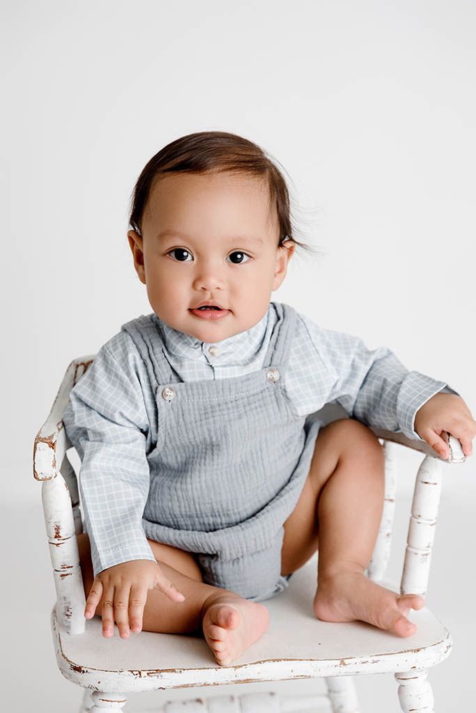 Adorable baby in gray overalls sitting on a vintage white wooden chair against a white background.