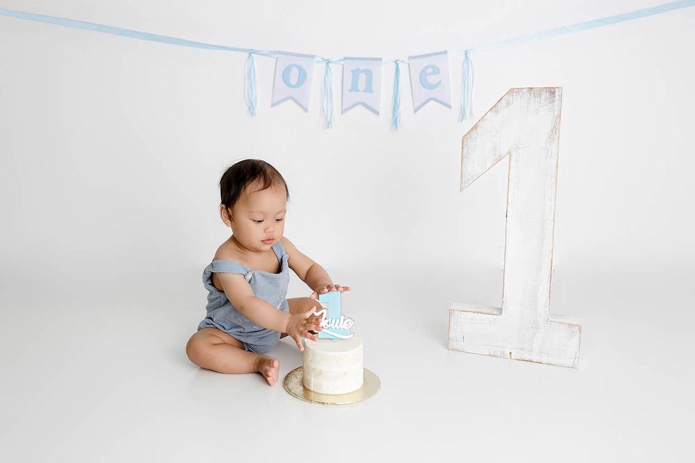 Baby boy in blue romper smashing his first birthday cake next to a wooden number one and banner.