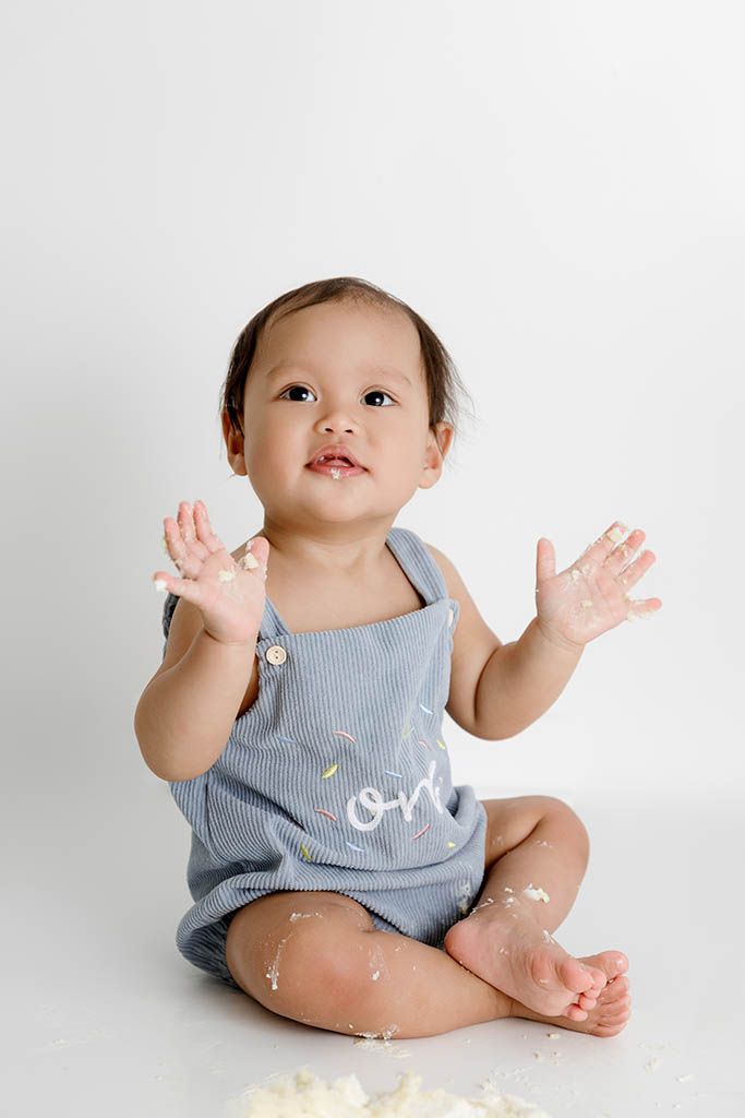 Cute baby in a blue romper sits on white floor with cake-covered hands raised during first birthday smash.