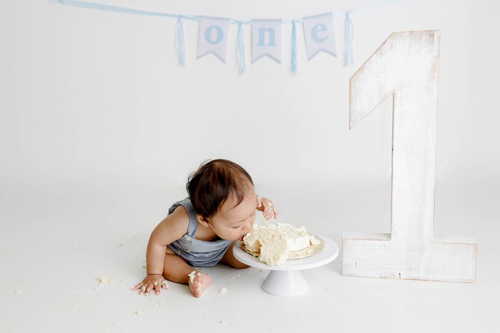 Baby boy smashing first birthday cake next to a number one prop with a blue 'one' banner above.