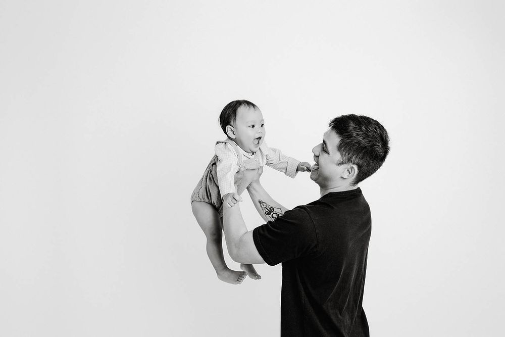 Black and white photo of a father lifting his smiling baby in the air against a white background.