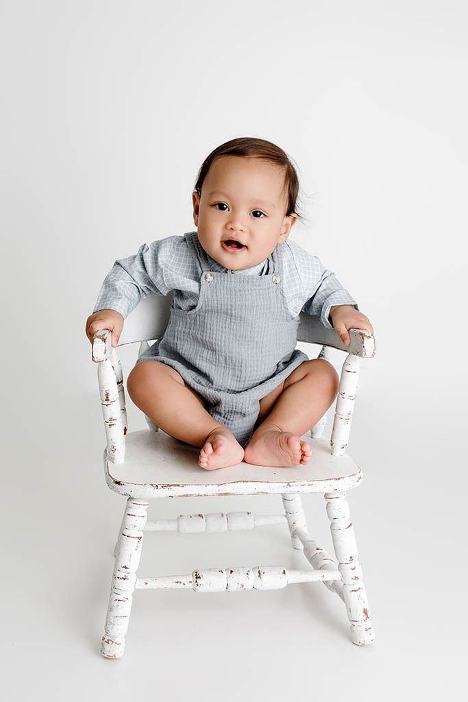 Smiling baby in blue romper sitting on a vintage white wooden chair against a white background.