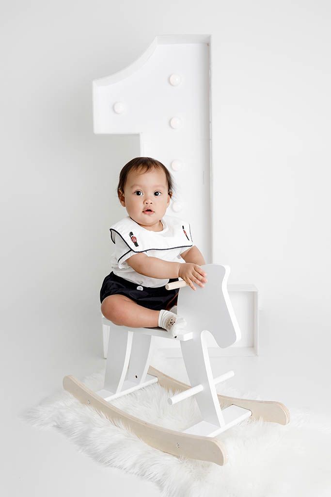 Baby in black and white outfit sits on a white rocking horse in front of a large number one prop.