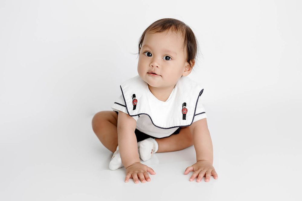 Adorable baby boy sitting in a white romper with toy soldier embroidery on a white background.