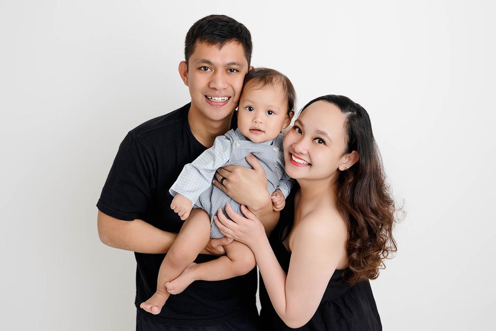 Happy Asian family portrait with smiling parents in black outfits holding their cute baby in blue romper.