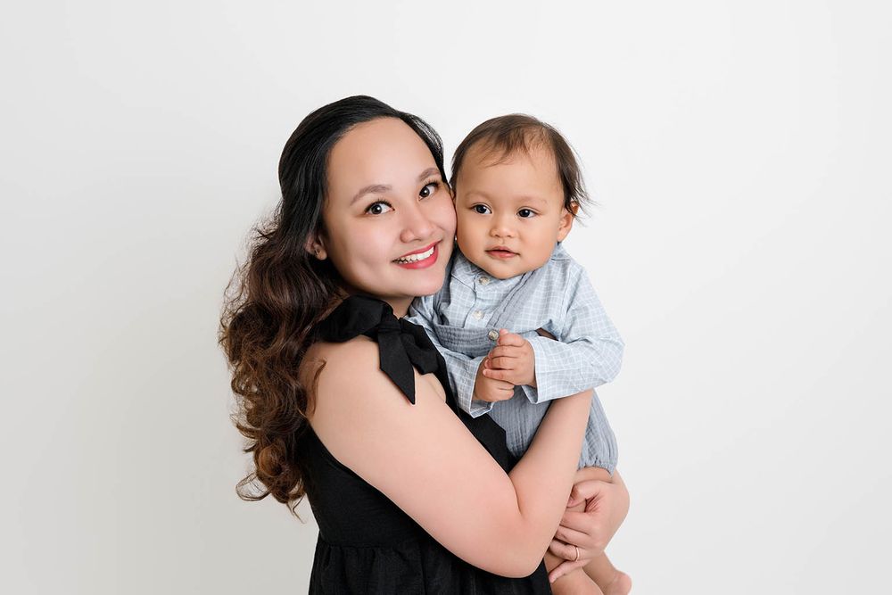 A smiling young woman in a black dress holds an adorable baby boy in a blue outfit against a white background.