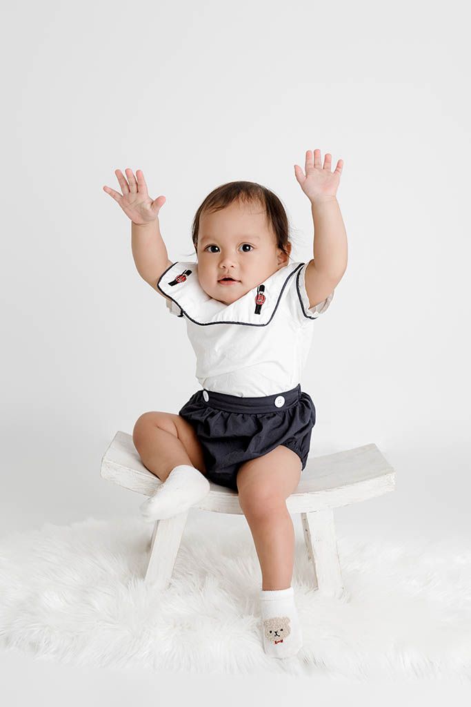 Adorable baby in navy sailor outfit sits on white bench with arms raised, wearing white socks on fluffy rug.
