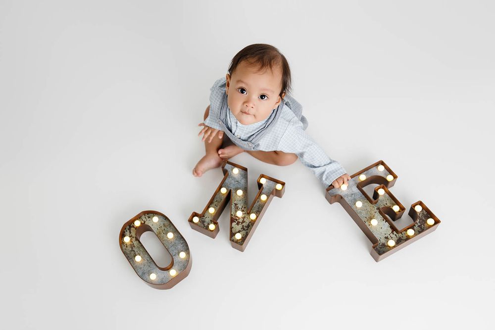 Baby boy in blue romper lying next to illuminated marquee letters spelling ONE for first birthday photo.