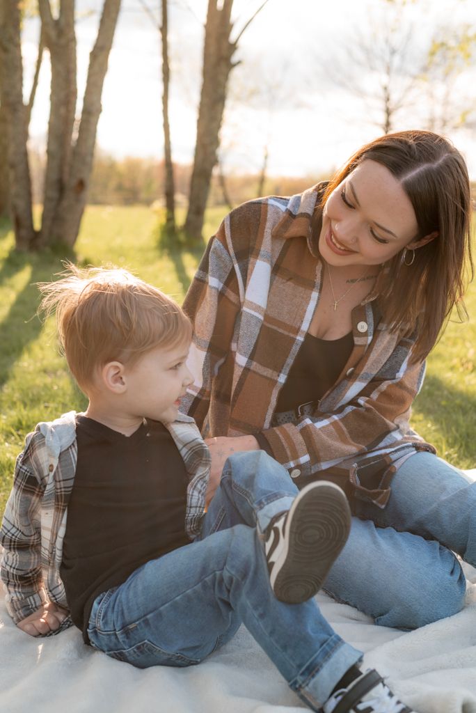 A smiling mother and young son sit on a blanket outdoors in golden sunlight, both wearing plaid shirts and jeans.
