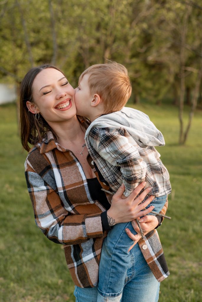 A smiling young mother in a plaid shirt holds her toddler son who gives her a sweet kiss on the cheek outdoors.