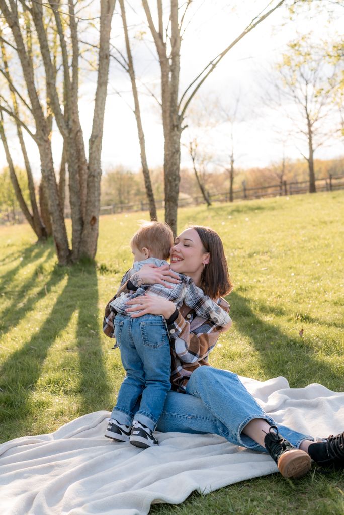 A young mother sits on a blanket in a sunny park, lovingly hugging her toddler in matching plaid shirts.