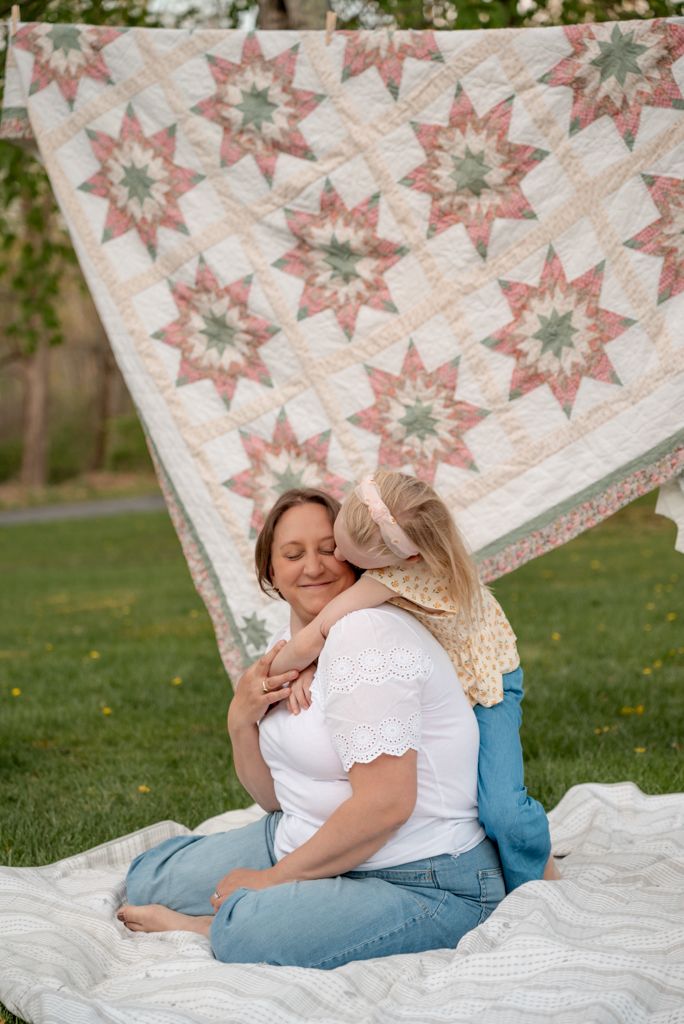 A mother and child share a tender hug on a blanket outdoors, with a colorful star quilt hanging behind them.