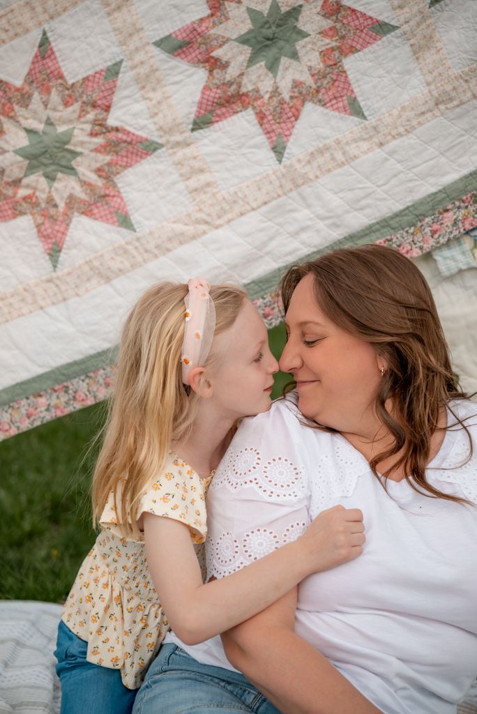 A young blonde girl and her pregnant mother share a sweet nose-to-nose moment outdoors against a colorful quilt backdrop.