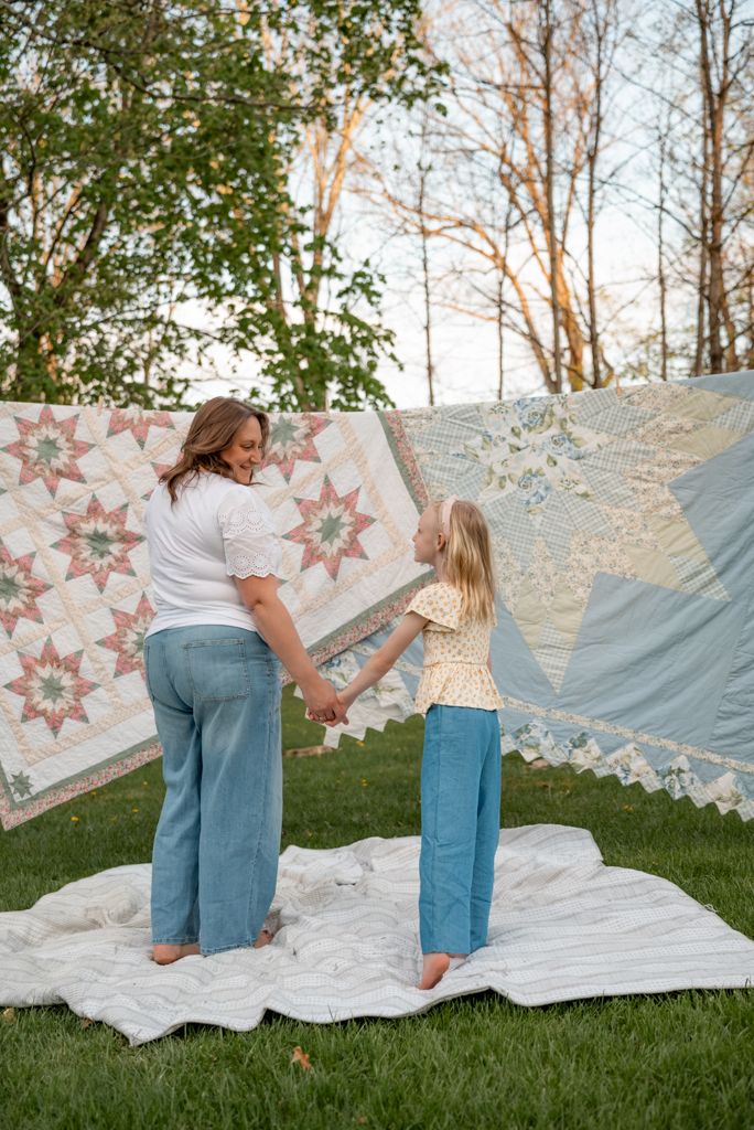 A mother and young daughter hold hands outdoors surrounded by colorful quilts hung on a line over green grass.