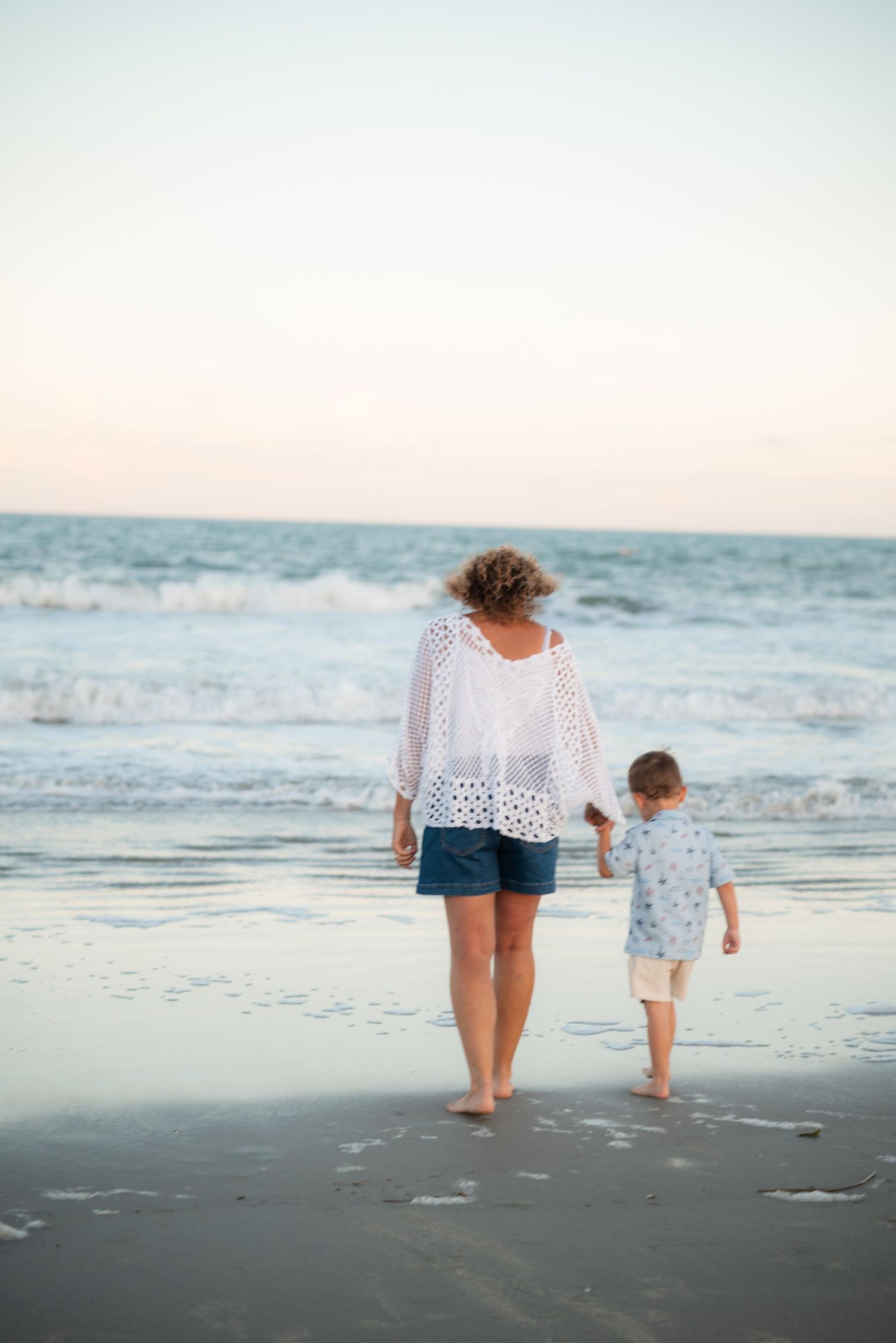 A woman and young child walk hand-in-hand along a sandy beach at dusk, waves gently rolling in behind them.