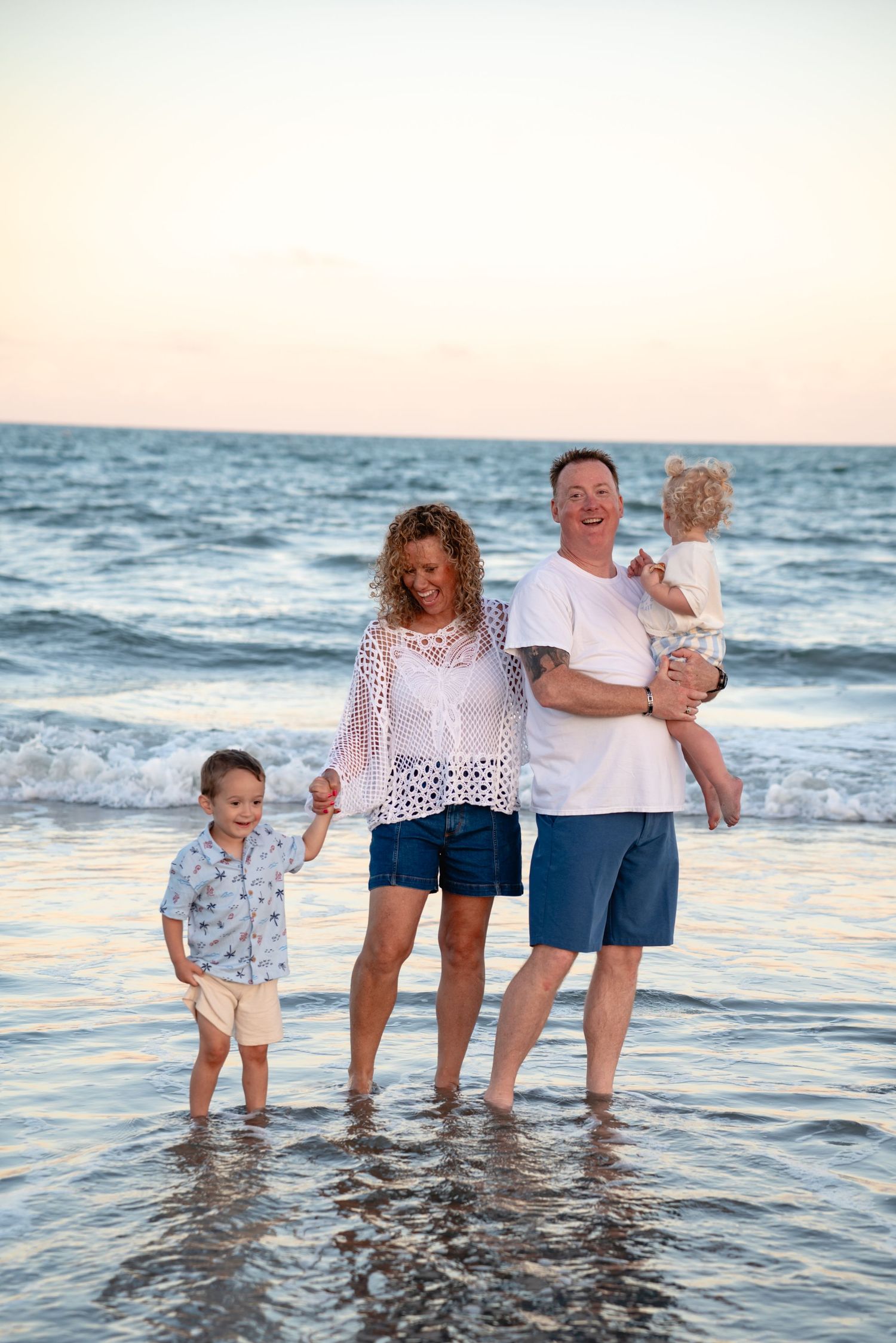 Family of four enjoying a beach sunset, parents with two young children playing in shallow ocean waves.