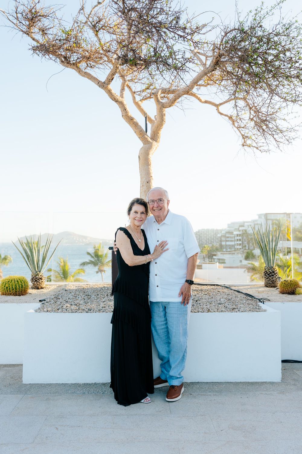 Elderly couple embracing and smiling outdoors near a desert tree and cacti with ocean views in background.