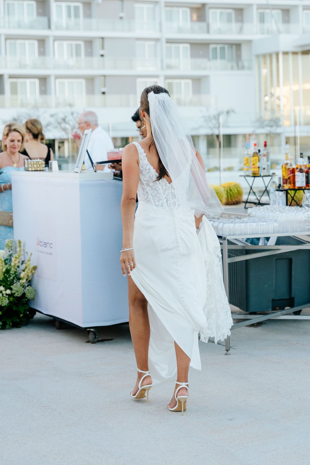 Bride in white lace gown and veil walks gracefully at outdoor wedding venue with bar setup in background.