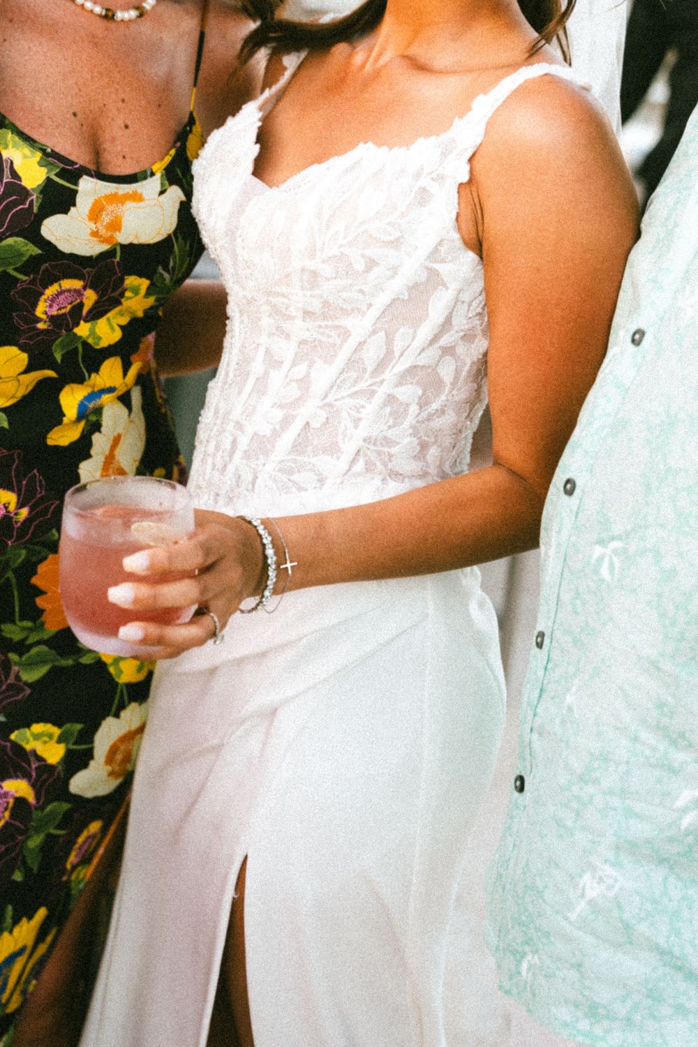 Woman in white lace dress holding a pink cocktail at a social gathering, flanked by guests in floral and blue attire.