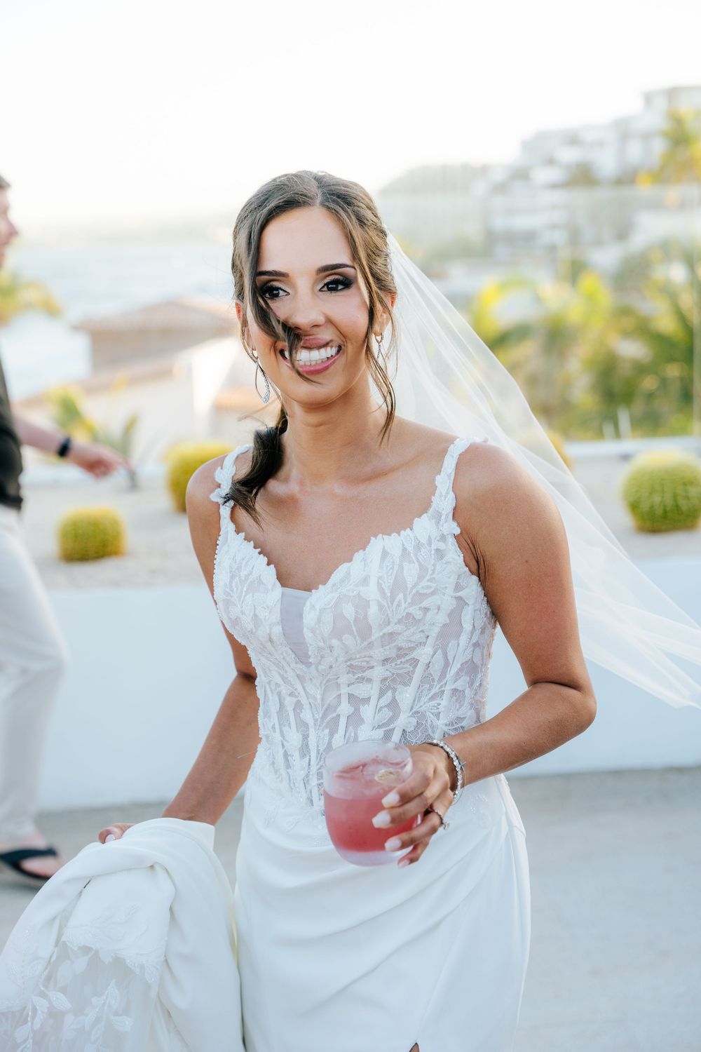 Smiling bride in white lace wedding gown and veil holding a pink cocktail at an outdoor wedding reception.