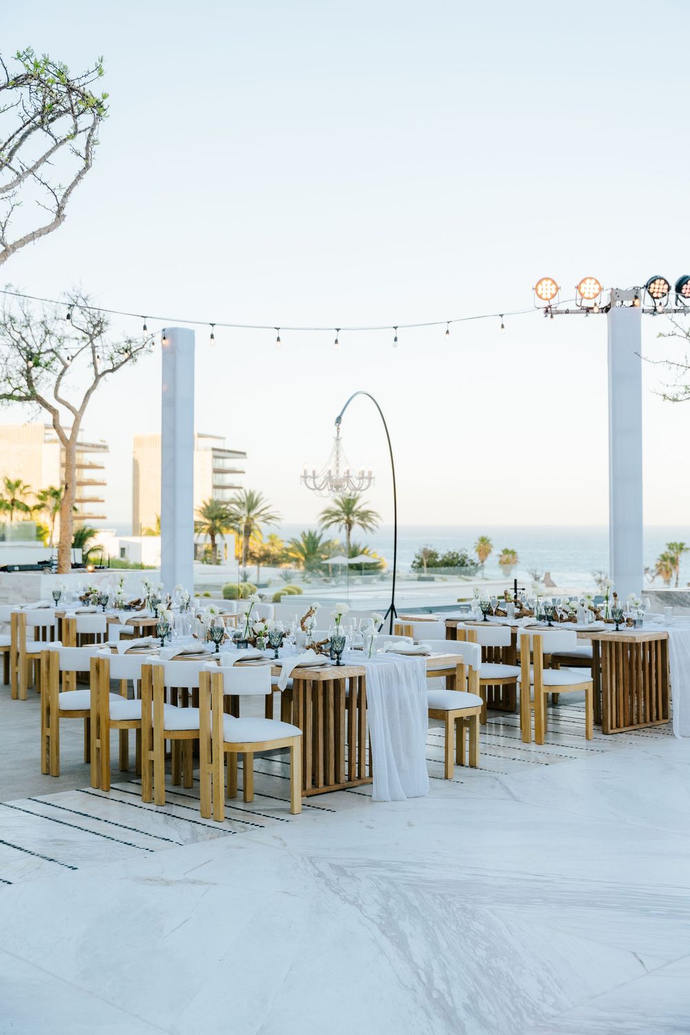 Elegant outdoor wedding reception setup with wooden chairs, white linens, string lights, and ocean views at dusk.