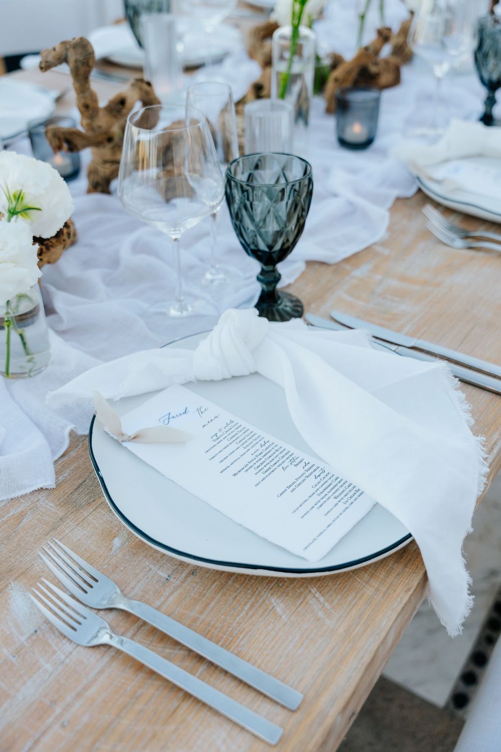 Elegant wedding table setting with white napkin, menu card, gray goblet, and wooden centerpiece on rustic table.