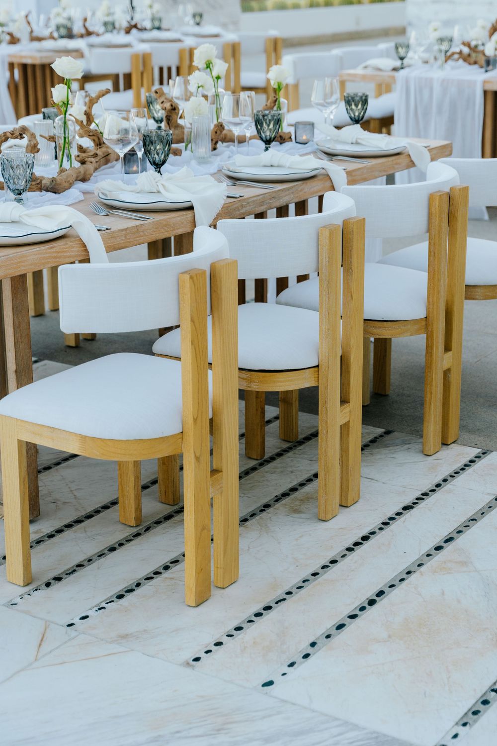 Elegant outdoor dining setup with wooden chairs, white cushions, and decorated tables with glassware and floral centerpieces.