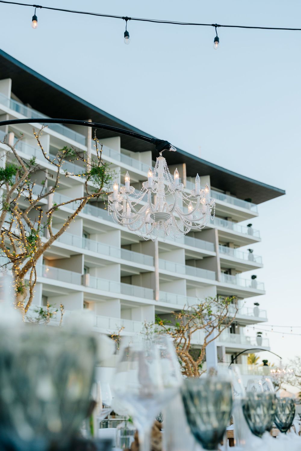 Elegant outdoor event setup with crystal chandelier, string lights, glassware, and a white multi-story hotel building backdrop.