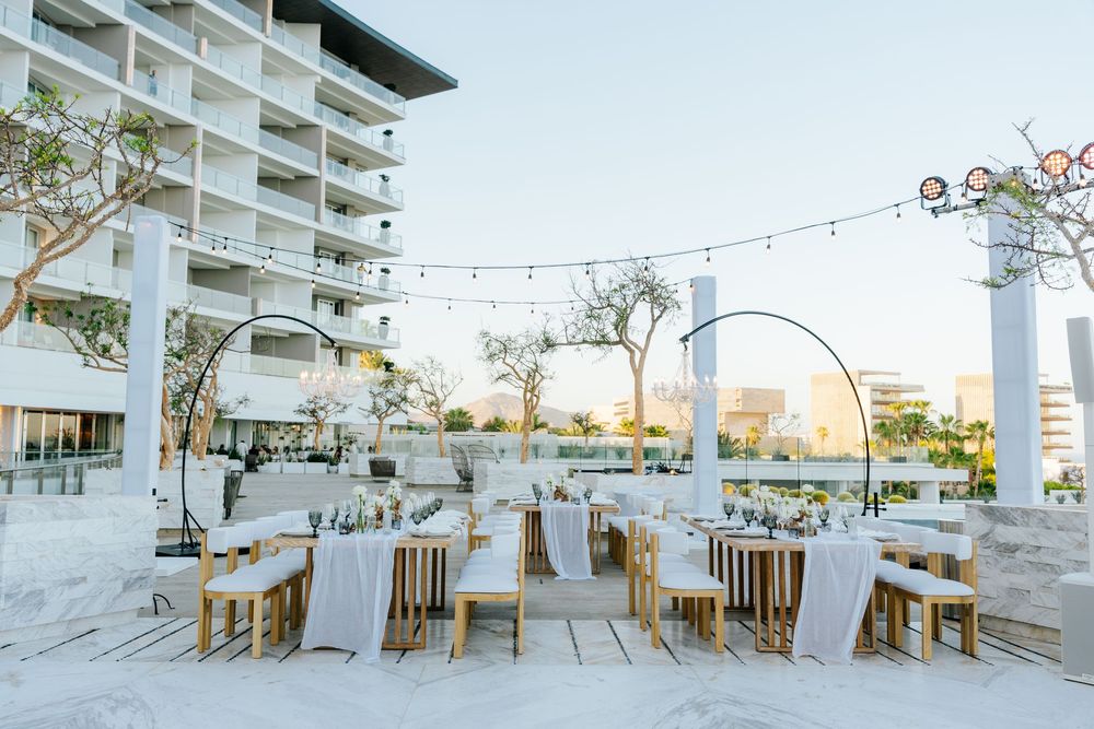 Elegant outdoor wedding reception with string lights, circular arches, and white-draped tables on a terrace near a hotel.