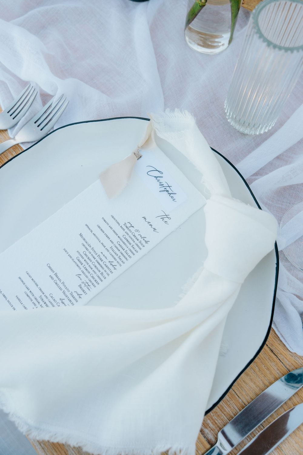 Elegant wedding place setting with white napkin, personalized menu card, and glassware on a wooden table.