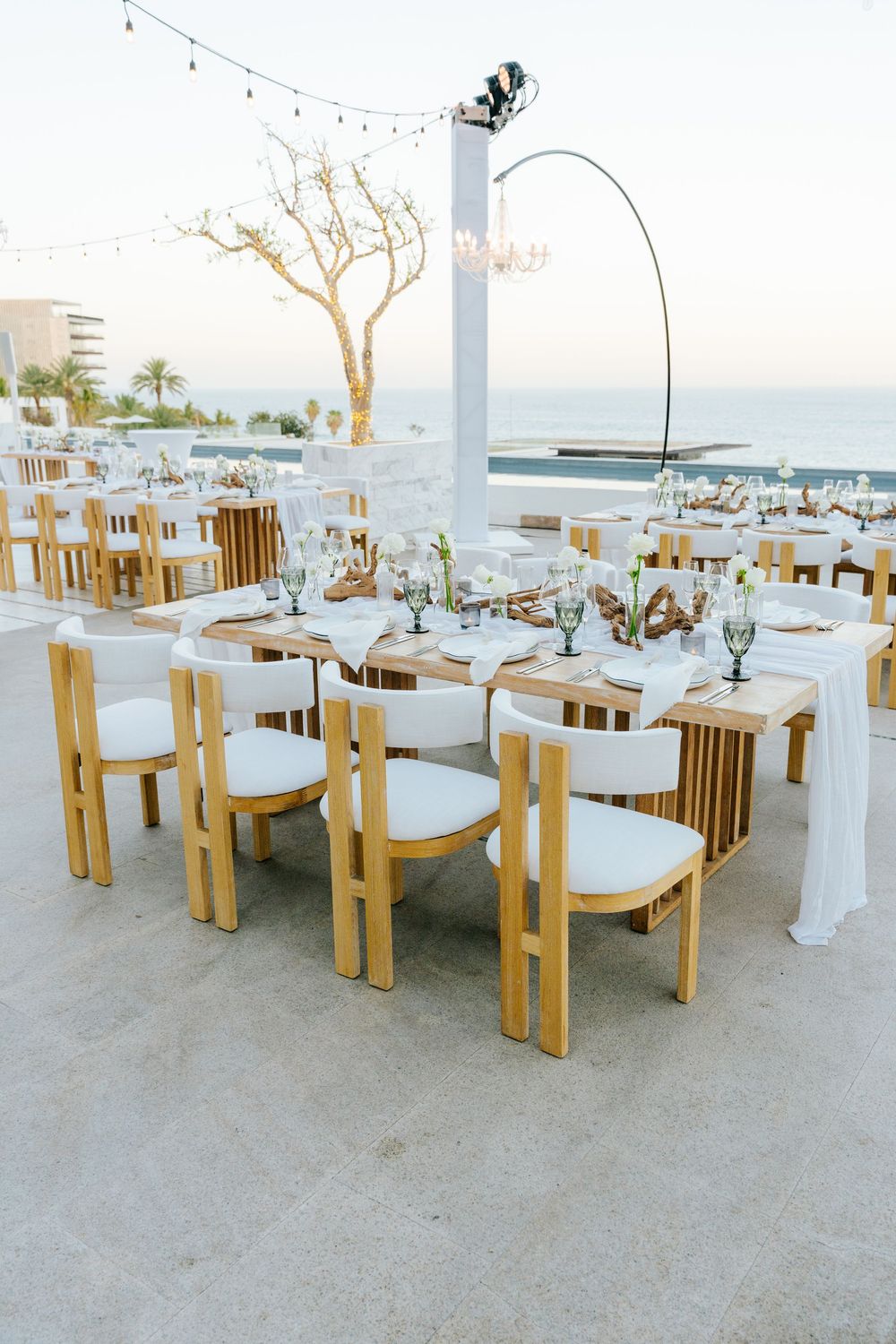Close-up of outdoor wedding reception tables with wooden chairs, white draping, glassware, and sea views at sunset.