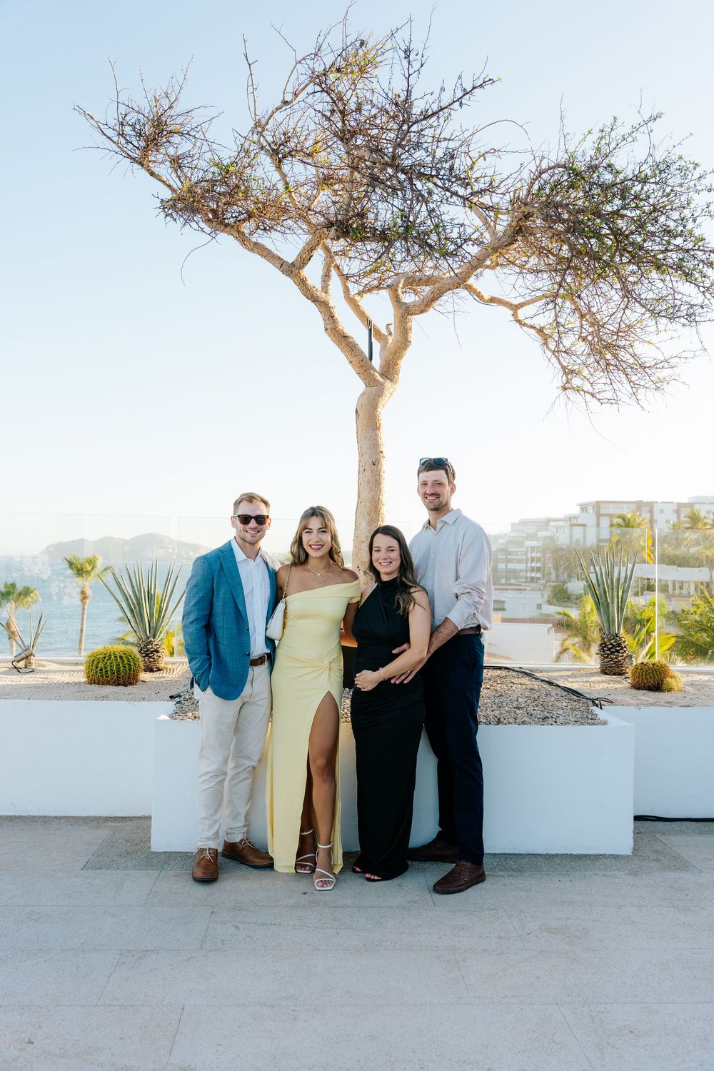 Two couples posing outdoors on a rooftop terrace with a bare tree backdrop and ocean views in Cabo.