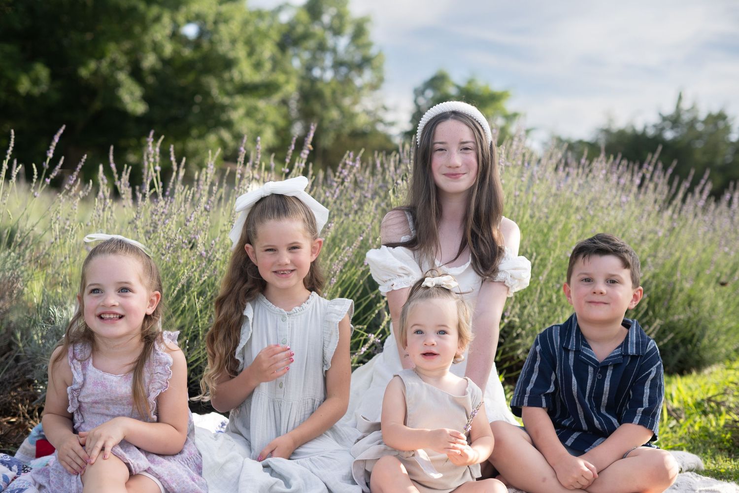 Five children sitting together outdoors in a lavender field on a blanket, smiling for a family portrait photo.
