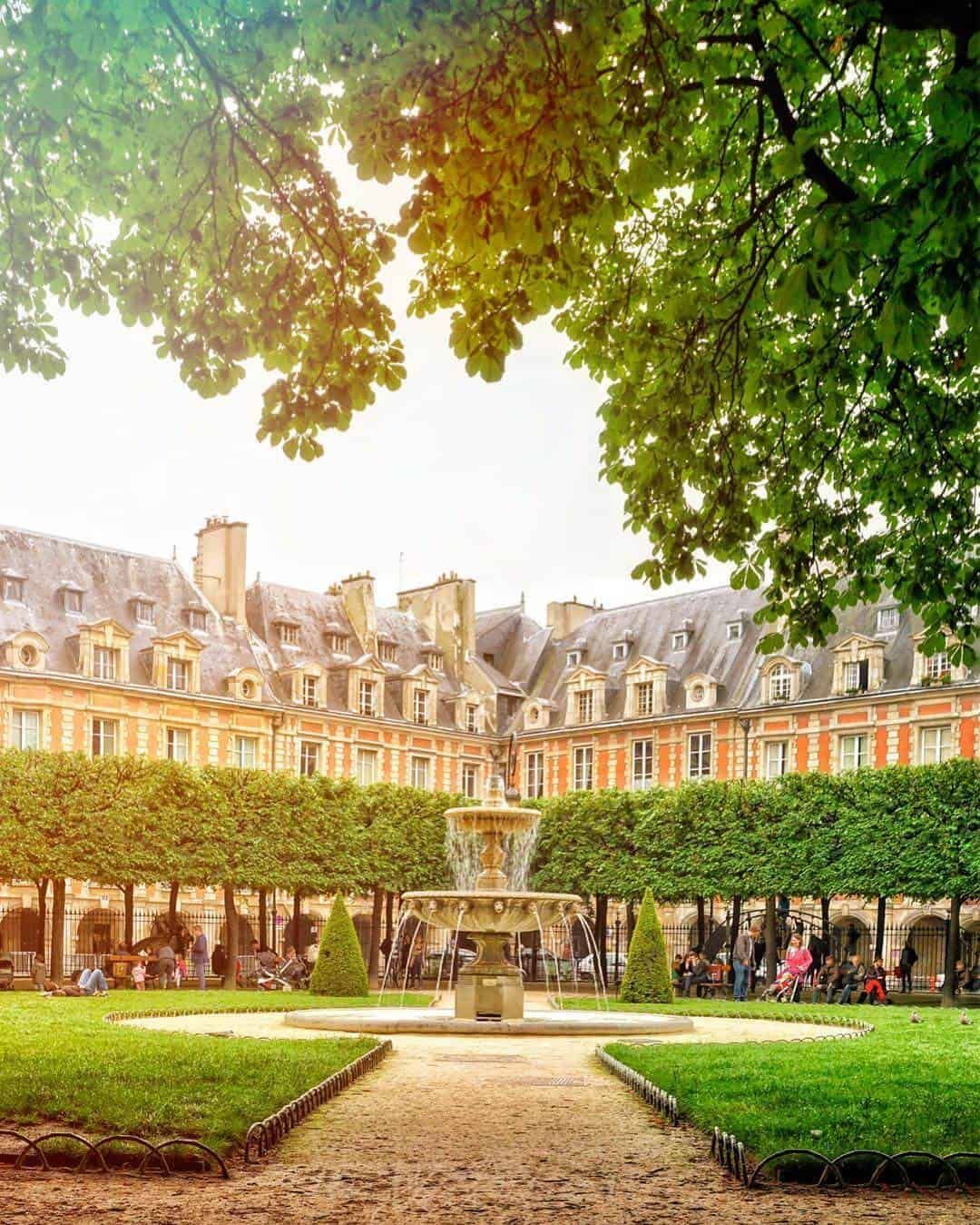 Place des Vosges garden in Paris with fountain, manicured hedges, and historic red brick buildings under lush green trees.