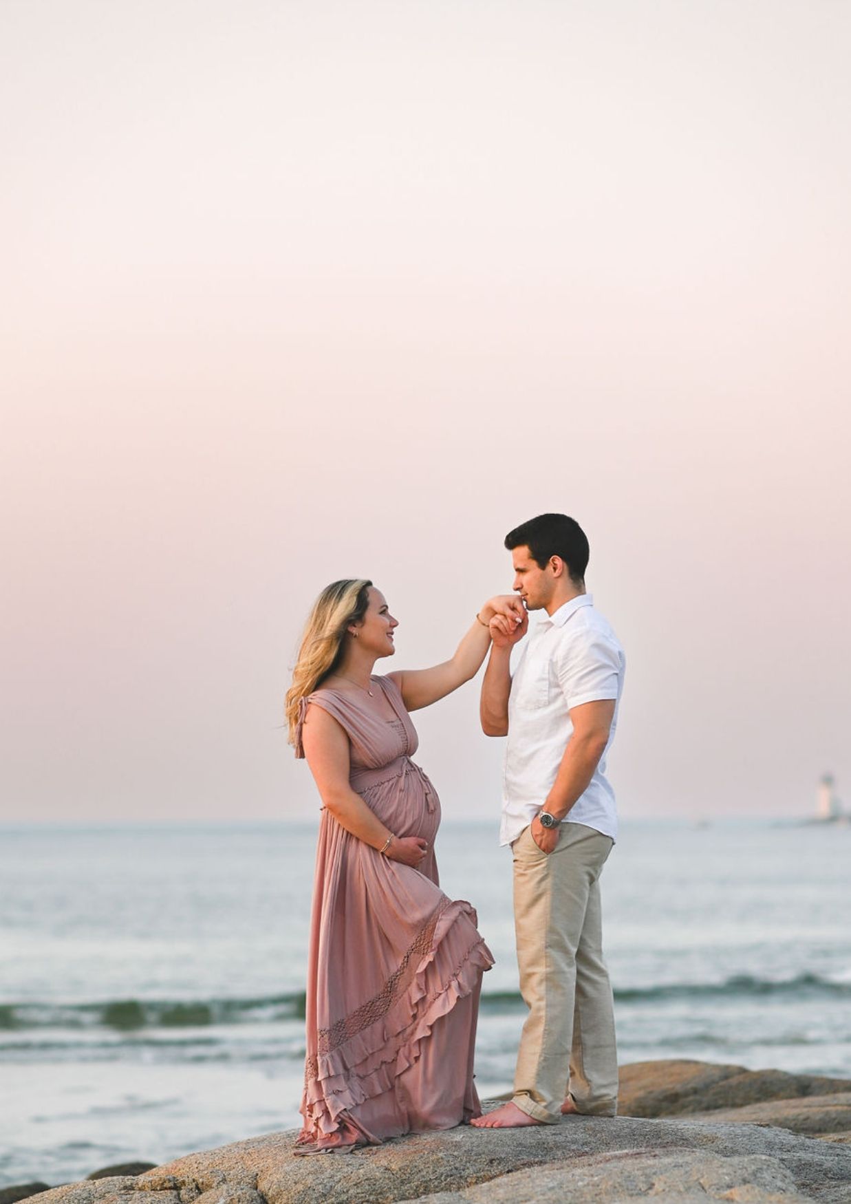 Pregnant woman in pink dress holds hands with man on rocky beach at sunset.