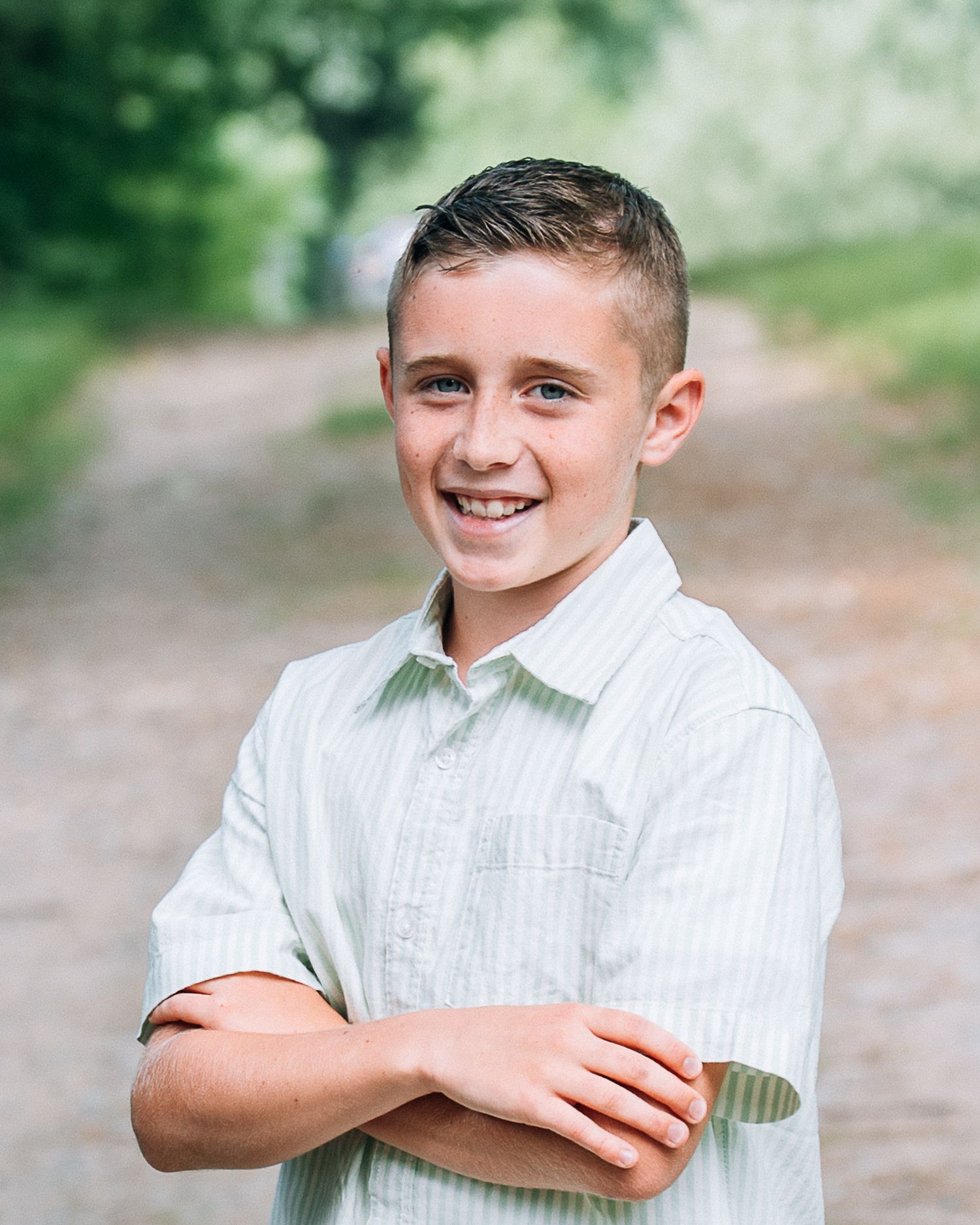 Smiling boy with arms crossed wearing a striped shirt, posing outdoors on a tree-lined path.