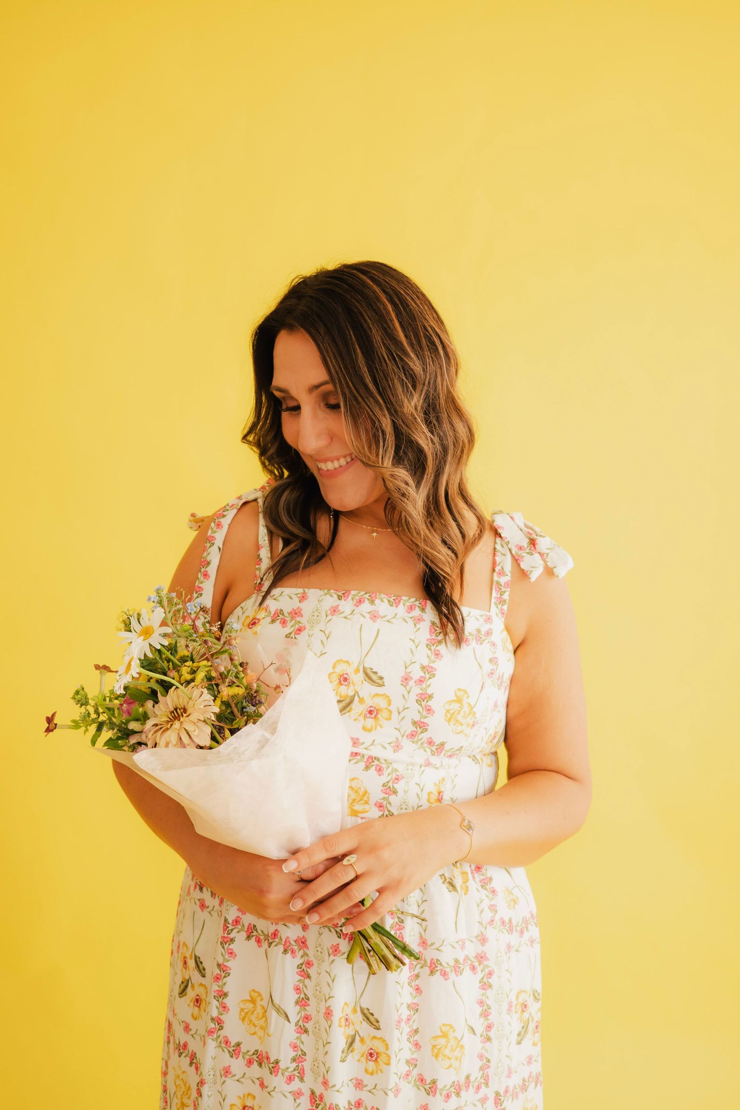 Smiling woman in floral dress holding a wildflower bouquet against a bright yellow background.
