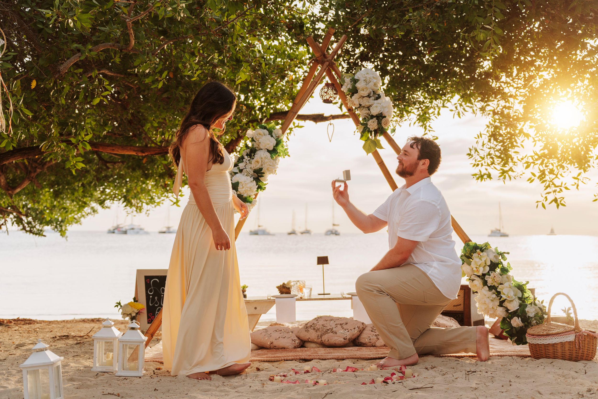 Couple holding hands at sunset beach proposal setup with floral arch, candles, and Marry Me sign on sandy shore.