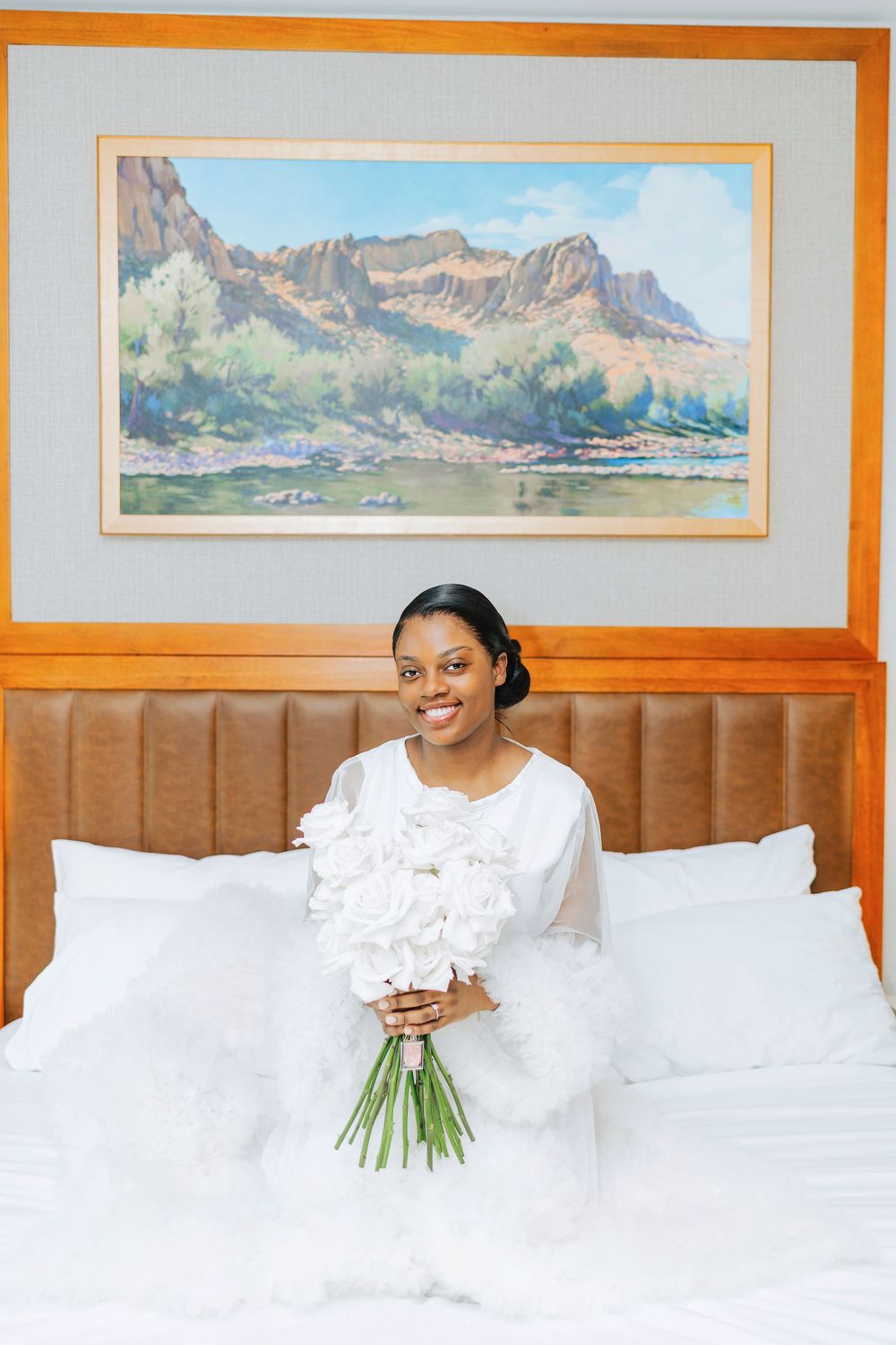 Smiling bride in white dress holding white peony bouquet on a hotel bed with a mountain landscape painting above.
