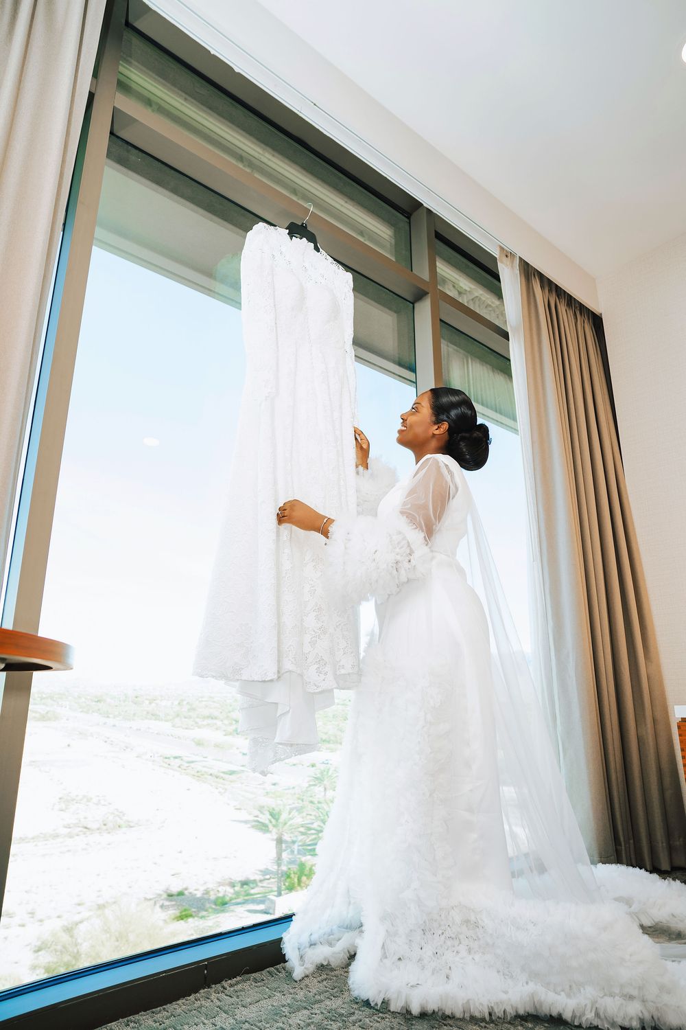 Bride in white lace robe admires her wedding dress hanging by a large window with a scenic view.