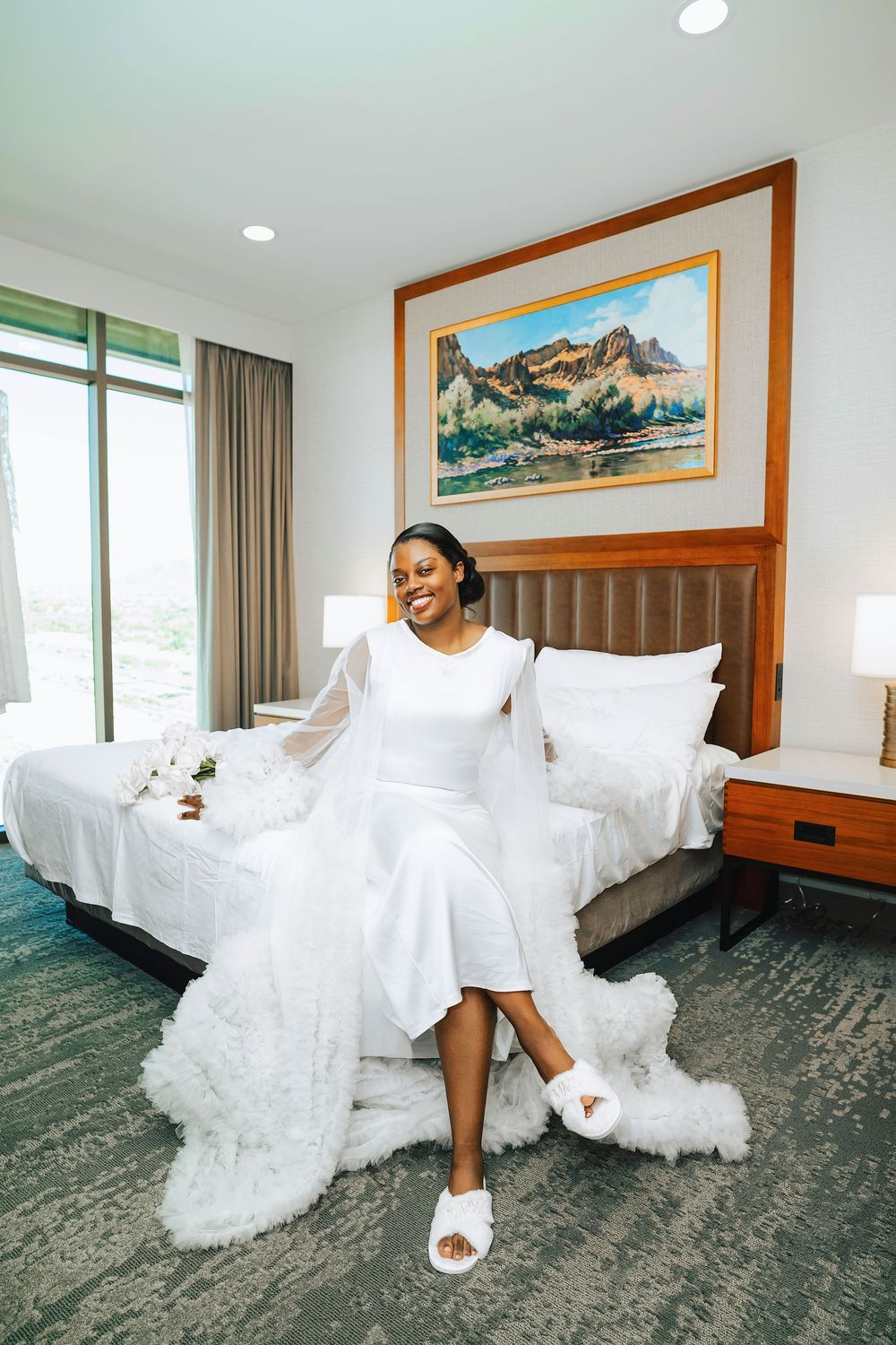 Bride in white wedding dress and fluffy slippers sits smiling on a hotel bed.