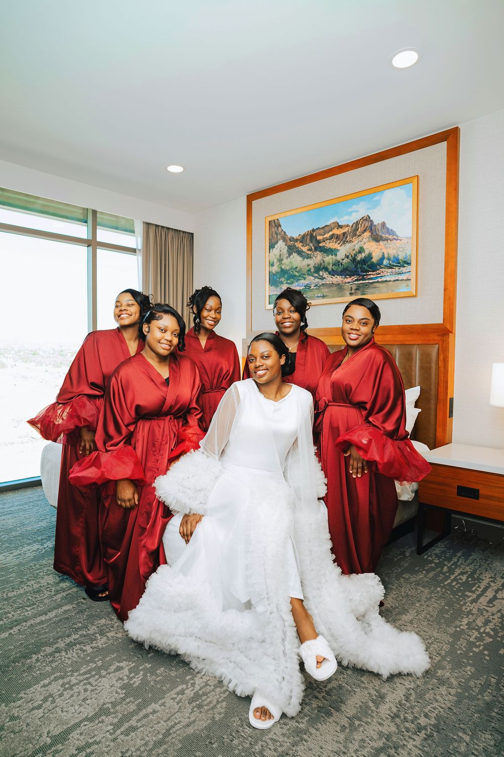 Bride in white wedding dress sits with five bridesmaids in red satin robes in a hotel room.