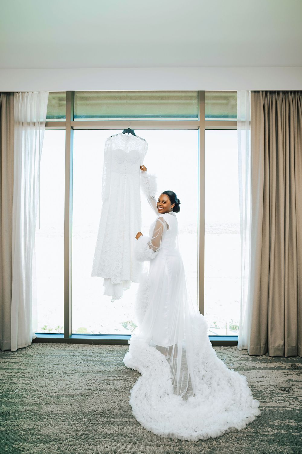 Joyful bride in lace wedding dress reaching up to touch her gown hanging by a large hotel window.
