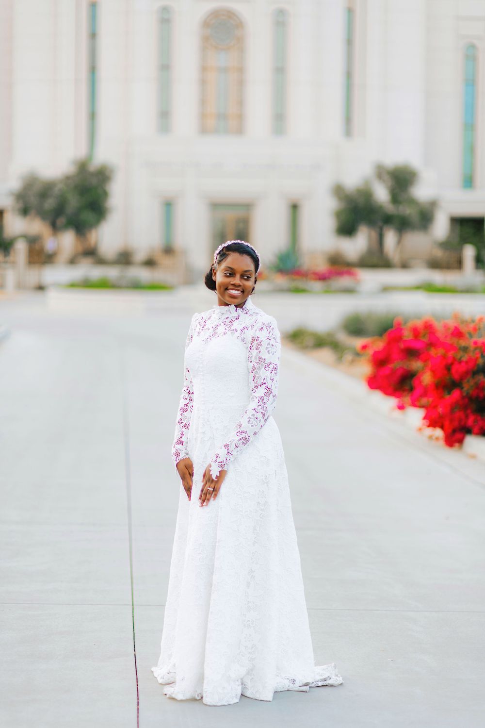 Smiling bride in white lace long-sleeve wedding gown standing outdoors near a white building with red flowers.