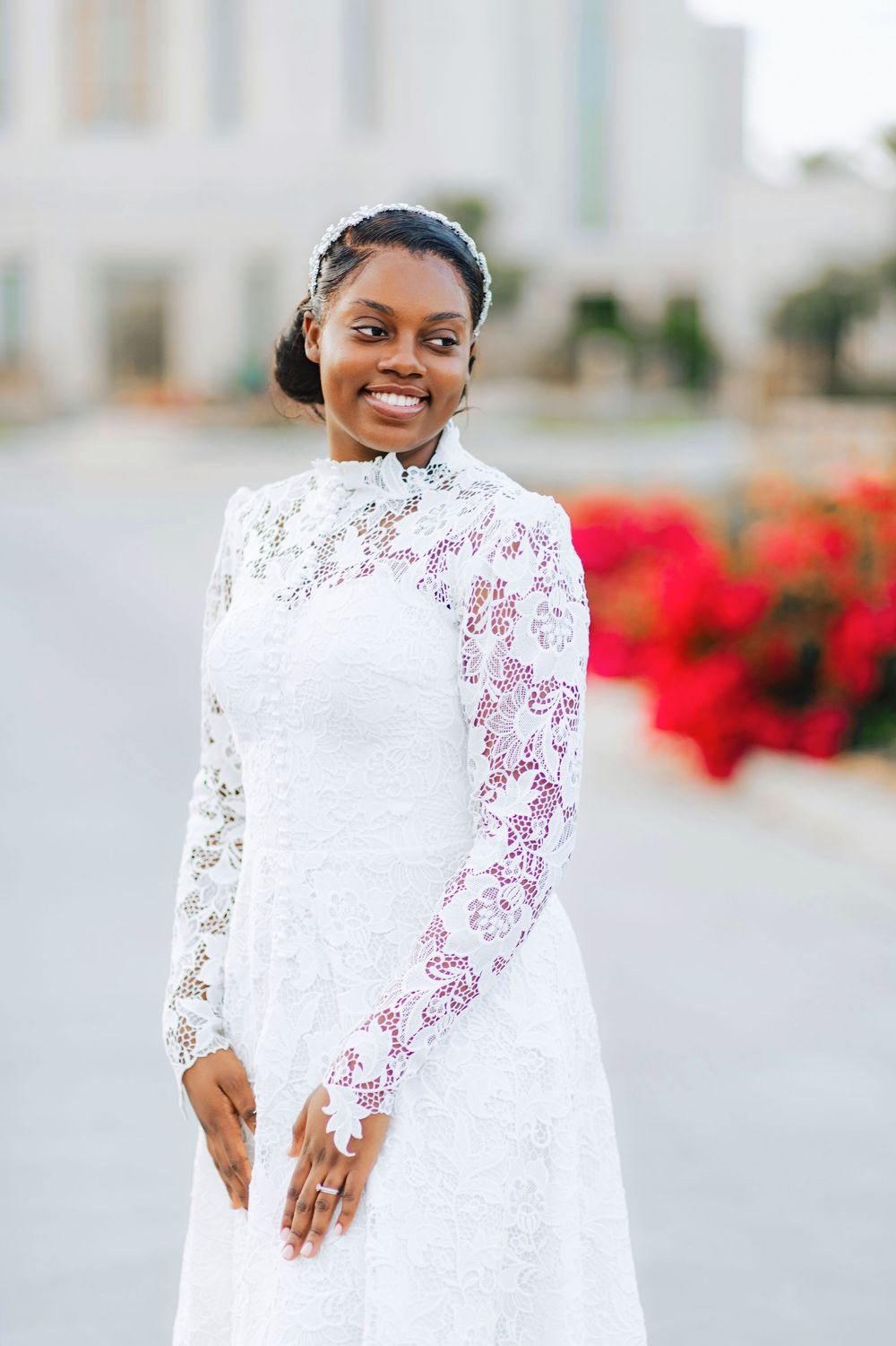 Smiling bride in elegant white lace long-sleeve gown posing outdoors with red flowers in background.
