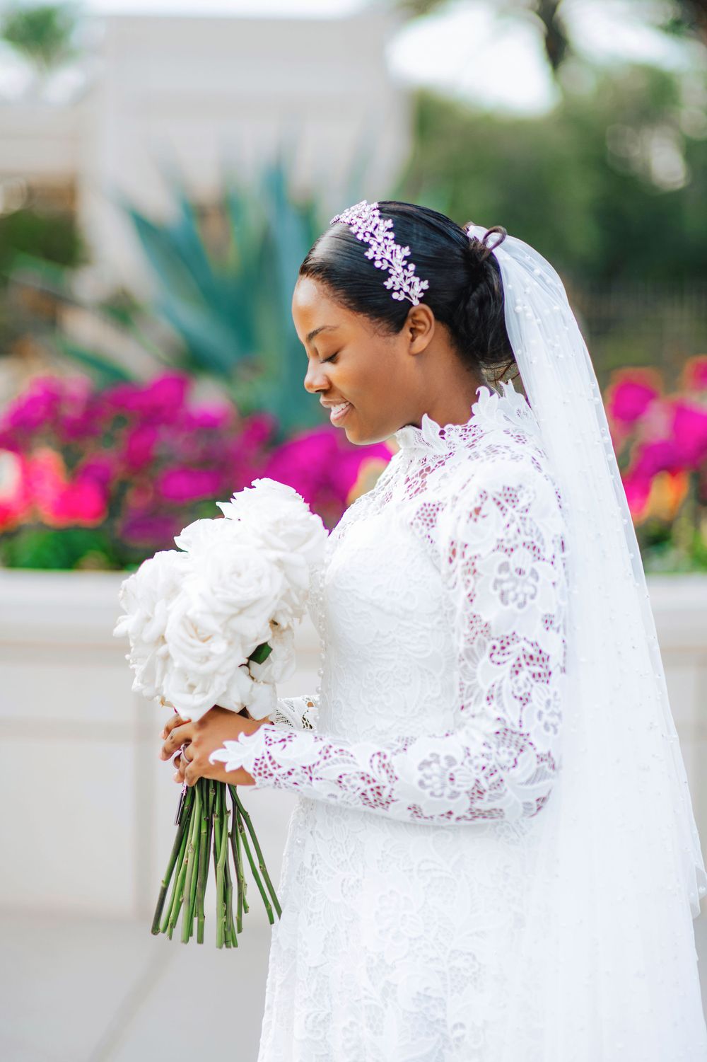 Bride in lace long-sleeve wedding dress holding white rose bouquet, wearing pink floral headpiece and veil outdoors.