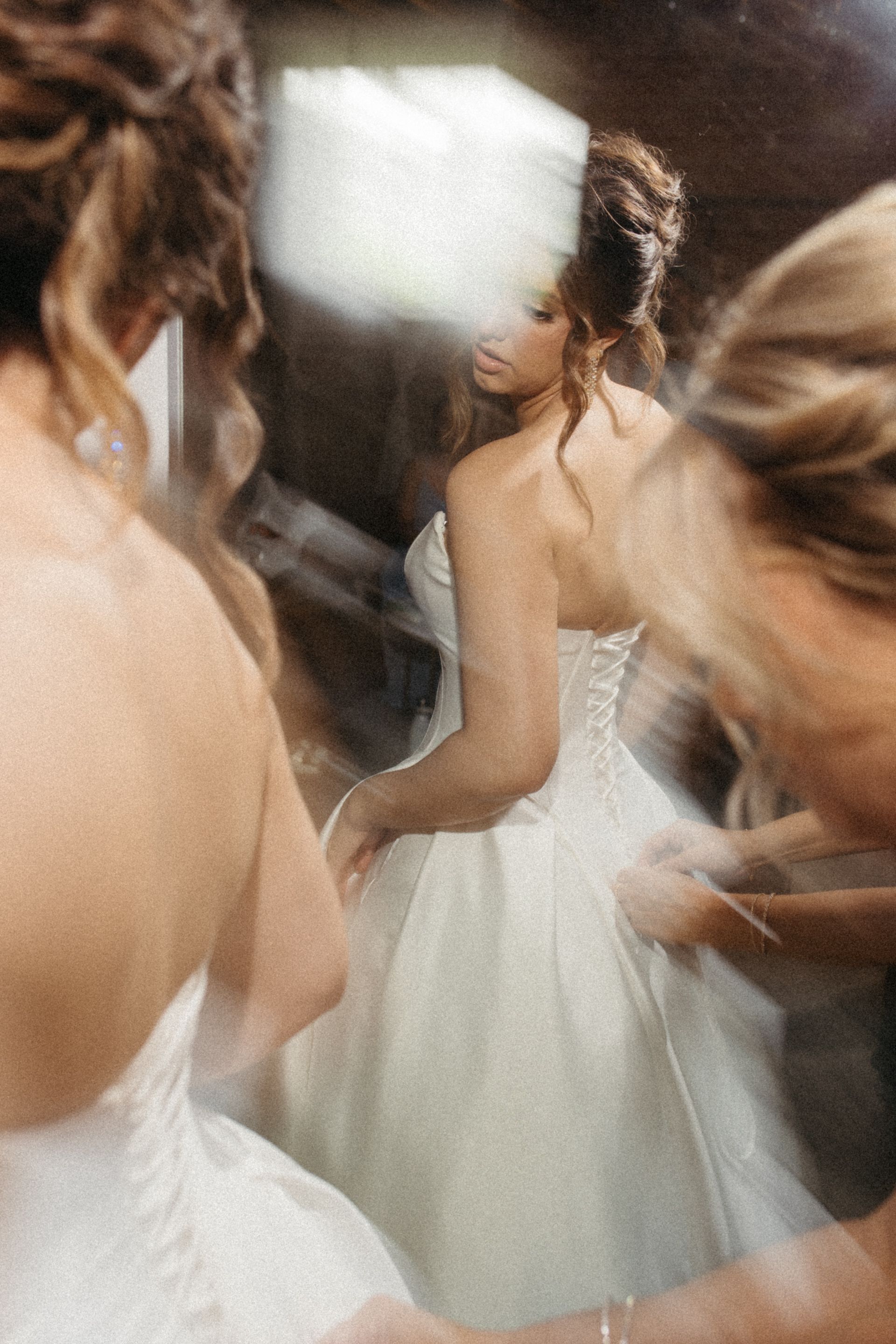 Bride in a white lace wedding gown being helped by bridesmaids, captured in a dreamy mirror reflection.