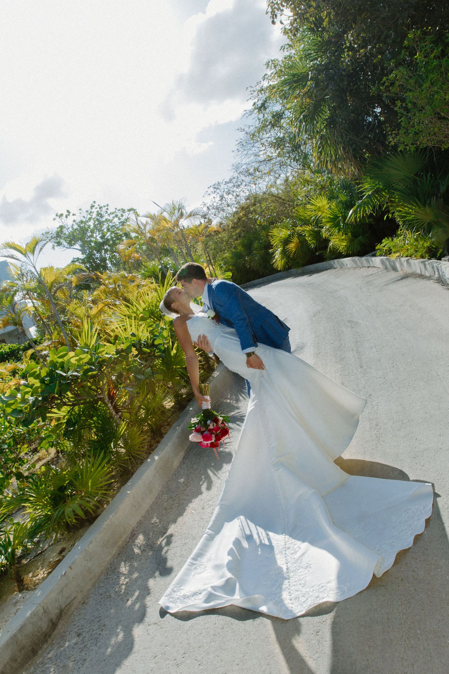 Bride and groom share a romantic dip kiss on a curved white pathway surrounded by lush tropical greenery.